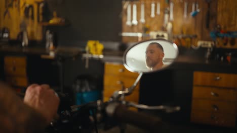 Confident-biker-starts-his-motorcycle-and-is-reflected-in-the-rear-view-mirror-in-his-garage-workshop