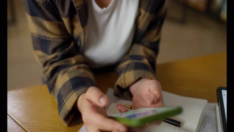 Close-up,-a-girl-in-a-white-T-shirt-and-a-plaid-shirt-sits-at-a-table-and-types-on-her-smartphone