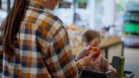 Close-up-a-mother-in-a-plaid-shirt-gives-an-apple-to-her-little-daughter-while-shopping-in-a-supermarket.-A-little-baby-girl-holds-an-apple-in-her-hands-and-examines-it-in-a-supermarket