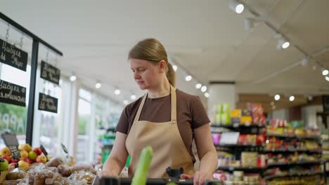 Confident-girl-in-a-brown-T-shirt-and-apron-puts-goods-on-the-counter-from-a-cart-during-her-work-in-a-supermarket