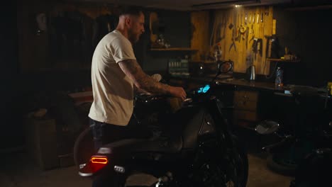 Confident-guy-in-a-white-t-shirt-with-a-tattoo-sits-on-his-bike-and-tests-it-in-his-garage-workshop