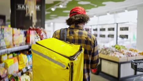 Rear-view-of-a-confident-brunette-guy-with-curly-hair-in-a-checkered-shirt-with-a-yellow-bag-a-food-delivery-man-walks-near-the-counter-in-a-supermarket-and-selects-the-necessary-products