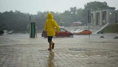 Happy-teenage-girl-in-a-yellow-jacket-and-orange-rubber-boots-runs-through-streams-of-water-and-puddles-during-heavy-rain-outdoors