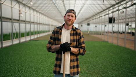 Portrait-of-a-confident-farmer-guy-in-a-cap-with-a-beard-in-a-plaid-shirt-who-poses-among-the-sprouts-of-young-plants-in-a-greenhouse-on-the-farm