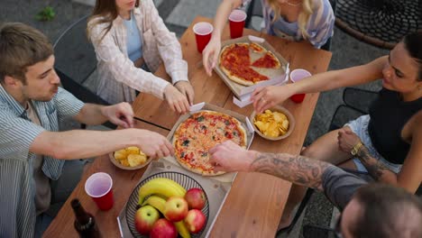 Top-view-of-a-happy-group-of-friends-sorting-out-pizza-from-paper-packaging-during-a-shared-lunch-at-a-table-in-the-courtyard-of-the-house