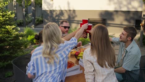 A-happy-company-holds-glasses-and-brown-bottles-in-their-hands-and-clinks-glasses-during-a-shared-lunch-at-a-table-in-the-courtyard-of-a-country-house-on-the-weekend