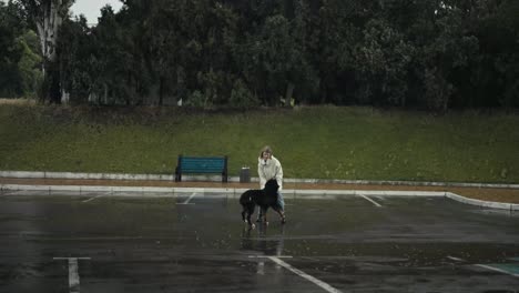 Glückliche-Blonde-Frau,-Die-Mit-Ihrem-Großen-Weißen-Hund-Davonläuft,-Während-Sie-Bei-Starkem-Regen-Im-Park-Spazieren-Geht