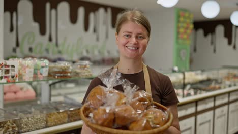 Portrait-of-a-happy-girl-in-a-brown-T-shirt-and-apron-holding-a-basket-of-buns-in-her-hands-while-working-in-the-confectionery-department-of-a-supermarket