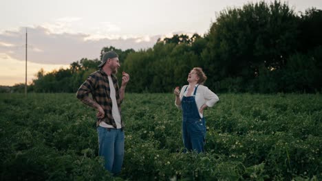 Un-Agricultor-Feliz-Con-Una-Camisa-A-Cuadros-Se-Comunica-Y-Ríe-Con-Su-Colega,-Una-Chica-Con-Un-Mono-De-Mezclilla-Come-Un-Tomate-Entre-Las-Plantas-Del-Campo