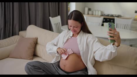 A-happy-pregnant-girl-is-vlogging-using-her-smartphone-about-her-pregnancy-and-expecting-a-baby-showing-little-socks-near-her-belly-on-the-sofa