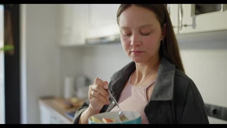 Pregnant-brunette-woman-in-a-gray-robe-stands-and-eats-fruit-salad-from-a-blue-bowl-in-the-kitchen-in-the-morning