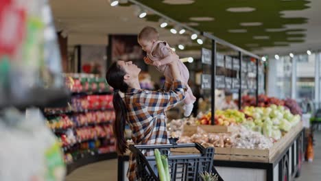 Happy-brunette-girl-in-a-plaid-shirt-holds-her-little-baby-in-her-arms-and-plays-with-him-during-her-shopping-in-the-supermarket