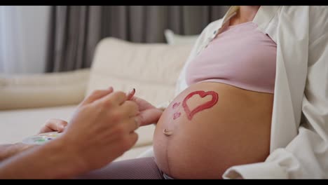 Close-up-a-man-using-red-paint-draws-a-heart-on-the-belly-of-his-pregnant-wife-on-the-sofa-at-home.-Happy-couple-expecting-baby-at-home