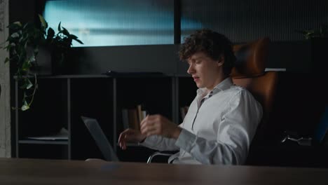 Side-view-of-a-tired-guy-with-curly-hair-in-a-shirt-getting-tired-and-touching-his-face-while-working-on-a-laptop-in-the-office