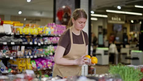 Una-Empleada-De-Supermercado-Con-Un-Delantal-Marrón-Claro-Coloca-Frutas-En-El-Mostrador-De-Un-Supermercado.-Una-Mujer-Con-Una-Camiseta-Marrón,-Una-Asistente-En-Un-Supermercado,-Examina-Frutas-En-El-Mostrador.