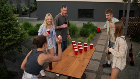 Ein-Selbstbewusstes-Brünettes-Mädchen-Wirft-Einen-Kleinen-Weißen-Ball-Und-Trifft-Einen-Becher-Bier-Während-Eines-Trinkspiels-Auf-Einer-Firmenfeier-Im-Innenhof-Eines-Landhauses