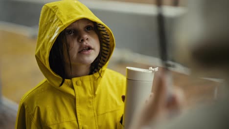 Over-the-shoulder-close-up-of-a-happy-little-teenage-girl-in-a-yellow-jacket-talking-to-her-mom-and-drinking-water-in-the-park-during-the-rain