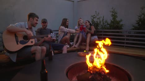 A-cheerful-group-of-friends-sing-listen-to-songs-on-the-guitar-and-communicate-while-spending-time-near-a-fire-in-the-courtyard-of-a-country-house