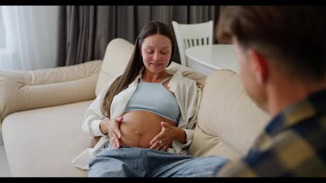 Over-the-shoulder-Happy-pregnant-brunette-woman-with-blue-eyes-lies-on-the-sofa-and-communicates-with-her-middle-aged-husband-at-home