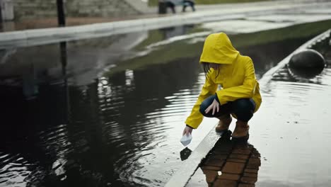 A-girl-in-a-yellow-jacket-floats-a-white-paper-boat-through-a-puddle-while-standing-on-the-curb-while-walking-in-the-drizzling-rain-on-the-street