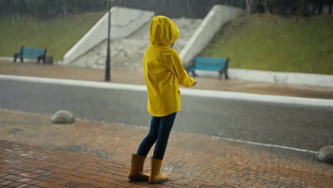 Rückansicht-Eines-Glücklichen-Teenager-Mädchens-In-Einer-Gelben-Jacke,-Das-Auf-Der-Veranda-Eines-Gebäudes-Steht-Und-Beim-Spaziergang-Im-Park-Auf-Den-Starken-Regen-Blickt