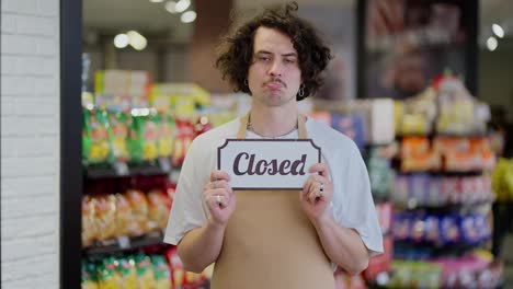 Portrait-of-a-sad-brunette-guy-in-an-apron-who-holds-in-his-hands-a-sign-with-the-inscription-closed-in-a-supermarket