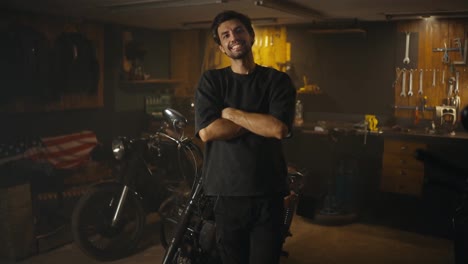 Portrait-of-a-happy-brunette-mechanic-guy-with-stubble-in-a-gray-T-shirt-who-poses-against-the-background-of-tools-and-motorcycles-in-his-workshop-studio