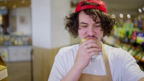 Close-up-of-a-confident-guy-with-curly-hair-and-a-red-cap-a-supermarket-worker-chews-a-bun-while-doing-inventory-on-the-shelves-in-the-supermarket