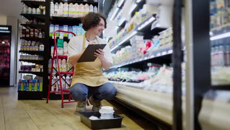 Ein-Glücklicher-Brünetter-Typ-Mit-Schnurrbart-In-Einer-Schürze-Verwendet-Ein-Tablet,-Um-Waren-In-Der-Milchabteilung-Eines-Supermarkts-Nachzuzählen