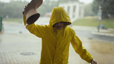 Cheerful-teenage-girl-in-a-yellow-jacket-with-a-hood-pours-water-from-a-rubber-boot-while-raining-and-walking-in-the-park