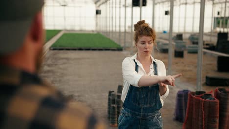 Over-her-shoulder-a-confident-girl-farmer-with-red-hair-in-a-white-shirt-shows-her-boyfriend-a-place-for-arranging-sprouts-of-young-plants-and-designing-a-greenhouse-on-the-farm