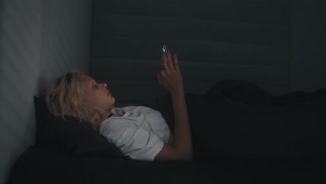 Blondes-Mädchen-In-Einem-Weißen-T-Shirt-Schaut-Auf-Das-Telefon,-Bevor-Es-Nachts-In-Einem-Grauen-Bett-Zu-Bett-Geht