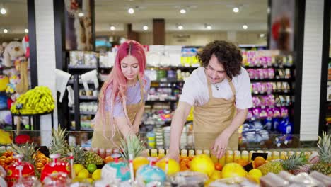 Ein-Brünetter-Typ-Mit-Lockigem-Haar-Und-Seine-Assistentin,-Ein-Mädchen-Mit-Rosa-Haaren-In-Einer-Gelben-Schürze,-Sortieren-Und-Inventarisieren-Produkte-In-Der-Zitrusfruchtabteilung-Eines-Supermarkts