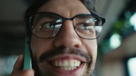 Close-up-of-a-happy-brunette-guy-with-stubble-in-glasses-communicates-on-a-green-smartphone-and-smiles-broadly-while-walking-around-the-office