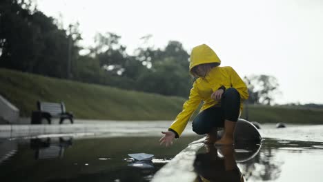 Una-Adolescente-Con-Una-Chaqueta-Amarilla-Hace-Flotar-Un-Barquito-De-Papel-En-Un-Charco-Cerca-De-La-Acera-En-Un-Parque.