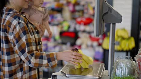 Close-up-shot-of-a-confident-mother-with-her-baby-in-her-arms-weighing-bananas-on-a-digital-clock-in-a-modern-supermarket