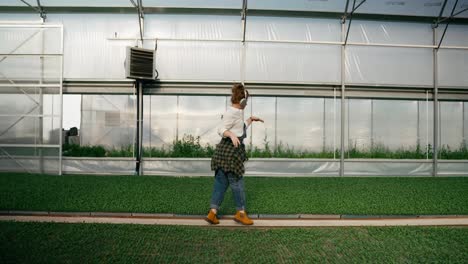 Vista-Lateral-De-Una-Mujer-Agricultora-Alegre-Y-Feliz-Escuchando-Música-Y-Bailando-Mientras-Camina-Junto-A-Las-Plántulas-Con-Plantas-En-Invernadero.