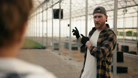 Over-the-shoulder-a-confident-guy-in-a-cap-with-a-beard-in-a-plaid-shirt-communicates-with-a-girl-with-red-hair-while-designing-a-greenhouse-and-working-on-a-farm