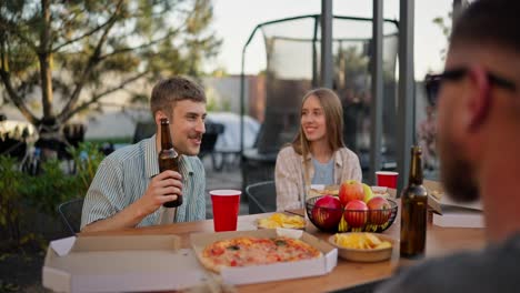 A-happy-blond-guy-with-a-bottle-of-beer-in-his-hands-communicates-with-his-friends-and-has-fun-during-lunch-in-the-courtyard-of-a-country-house-at-the-table