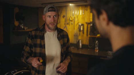 Over-his-shoulder-a-confident-mechanic-in-a-plaid-shirt-gives-advice-to-a-young-trainee-about-motorcycle-repairs-during-a-casual-conversation-in-a-workshop