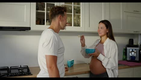 Happy-pregnant-brunette-woman-feeding-her-husband-fruit-salad-using-a-fork-in-the-kitchen-in-the-morning