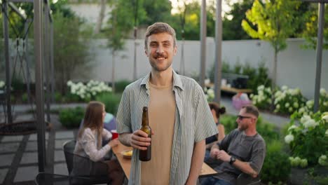 Portrait-of-a-happy-blond-guy-with-a-mustache-with-a-bottle-of-brown-beer-in-his-hands-while-relaxing-with-a-company-in-the-backyard-of-a-country-house