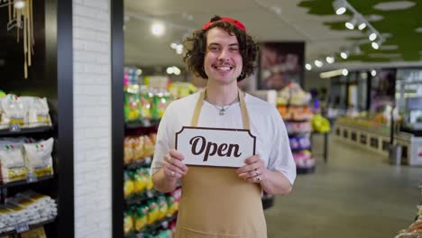 A-happy-brunette-guy-with-curly-hair-and-a-yellow-apron-holds-in-his-hands-a-sign-with-the-inscription-Open-in-the-supermarket