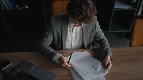 Top-view-of-a-confident-young-guy-in-a-gray-jacket-examining-charts-and-documents-work-reports-while-sitting-at-a-table-in-the-office