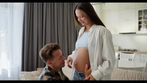 Happy-middle-aged-man-sits-down-and-kisses-his-pregnant-woman-belly-after-which-he-listens-to-the-child-at-home-in-a-modern-apartment