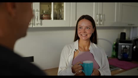 Over-the-shoulder-a-happy-pregnant-brunette-girl-in-a-white-shirt-drinks-tea-from-a-blue-cup-and-talks-with-her-husband-in-the-morning-in-the-kitchen