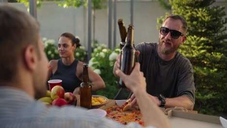 Por-Encima-Del-Hombro,-Un-Chico-Moreno-Y-Feliz-Con-Gafas-De-Sol-Choca-Una-Botella-De-Cerveza-Marrón-Con-Su-Amigo-Durante-El-Almuerzo-En-El-Patio-De-Una-Casa-De-Campo.