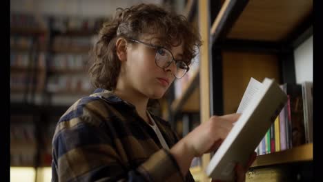 Confident-girl-with-curly-hair-wearing-glasses-reads-a-book-near-the-shelves-in-a-library-with-a-lot-of-books