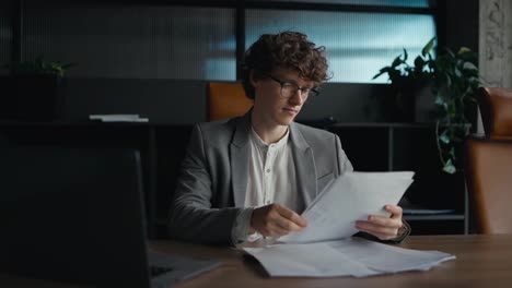 Confident-young-guy-with-curly-hair-in-a-gray-jacket-examines-papers-and-leans-back-in-his-chair-to-relax-while-working-in-the-office