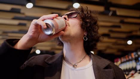 A-confident-and-cool-brunette-guy-with-curly-hair-and-black-sunglasses-walks-through-a-supermarket-and-drinks-lemonade-during-his-shopping